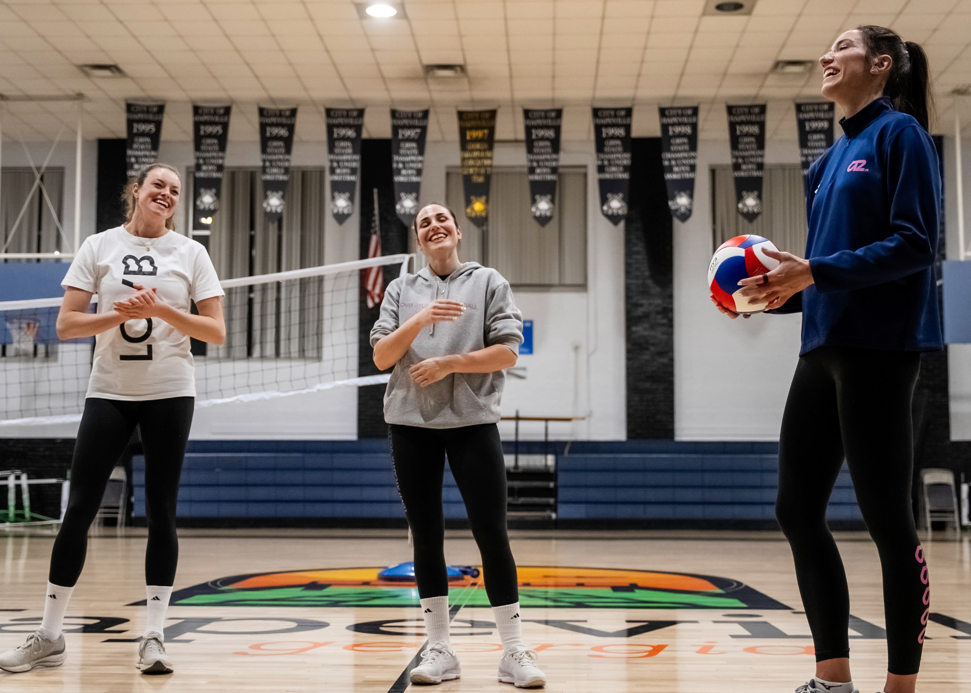Image of three women smiling in a gym with a volleyball net, one holding a volleyball.